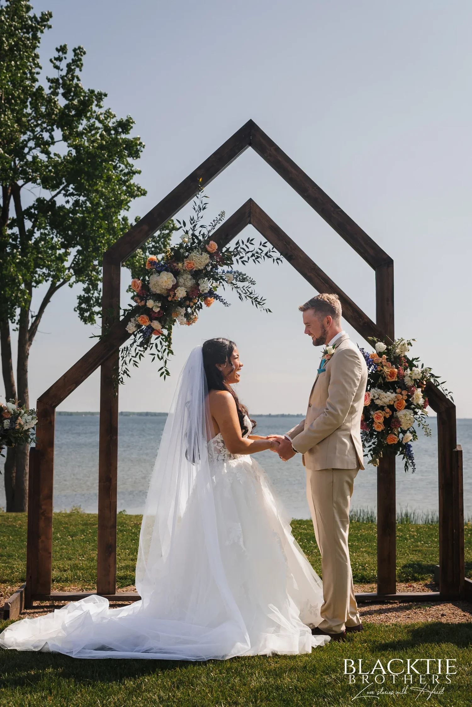 Carolina and Josh holding hands under the wooden double arch decorated with flowers, the lake behind them