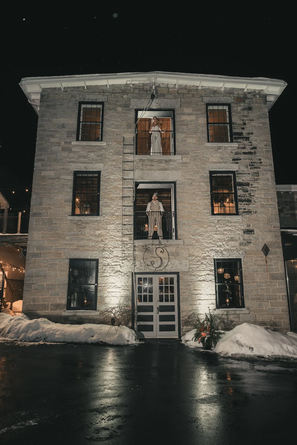 The illuminated stone facade of The Sinclair of Skaneateles, with Jill and Nick framed in its upper windows on their December wedding night