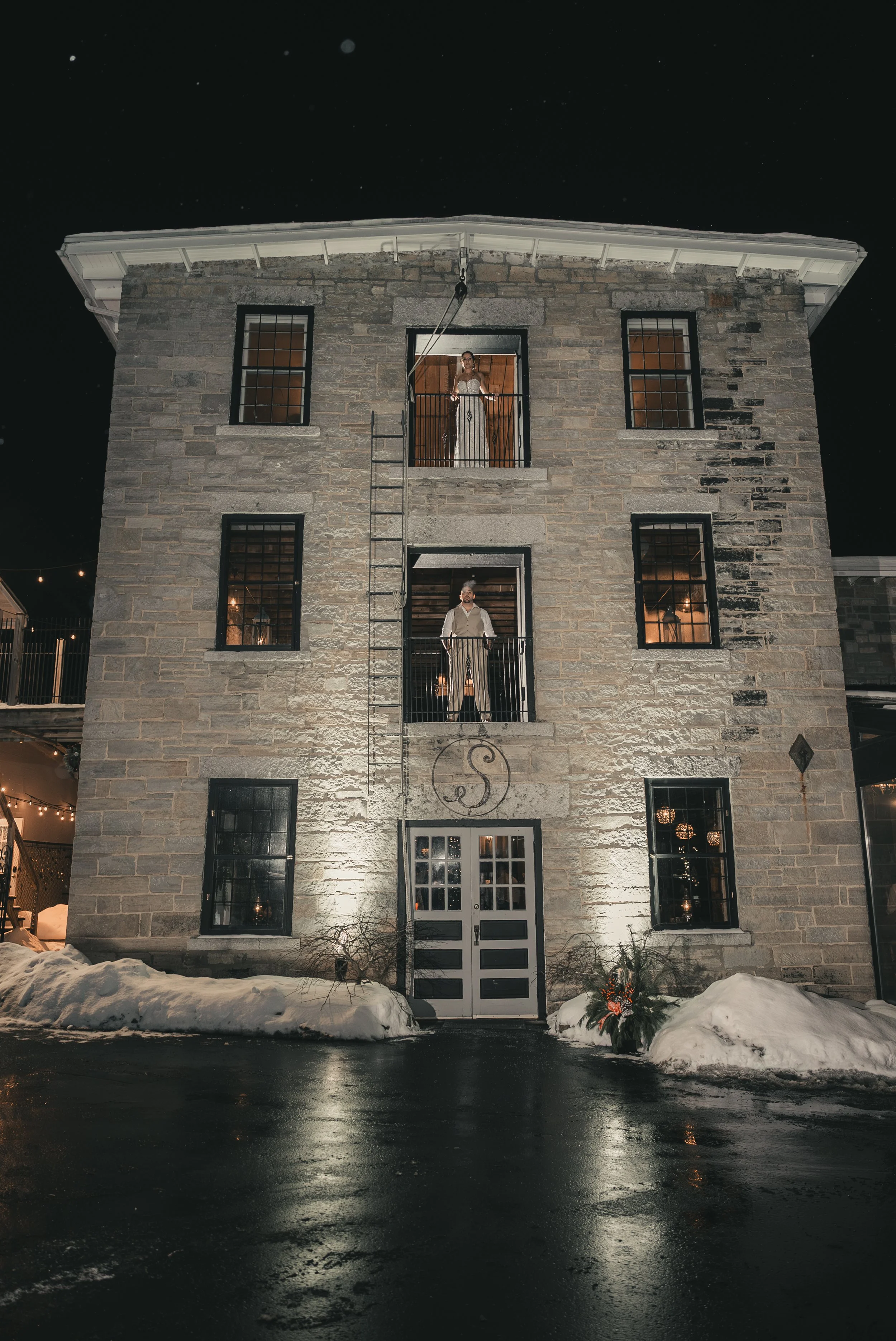 The illuminated stone facade of The Sinclair of Skaneateles, with Jill and Nick framed in its upper windows on their December wedding night