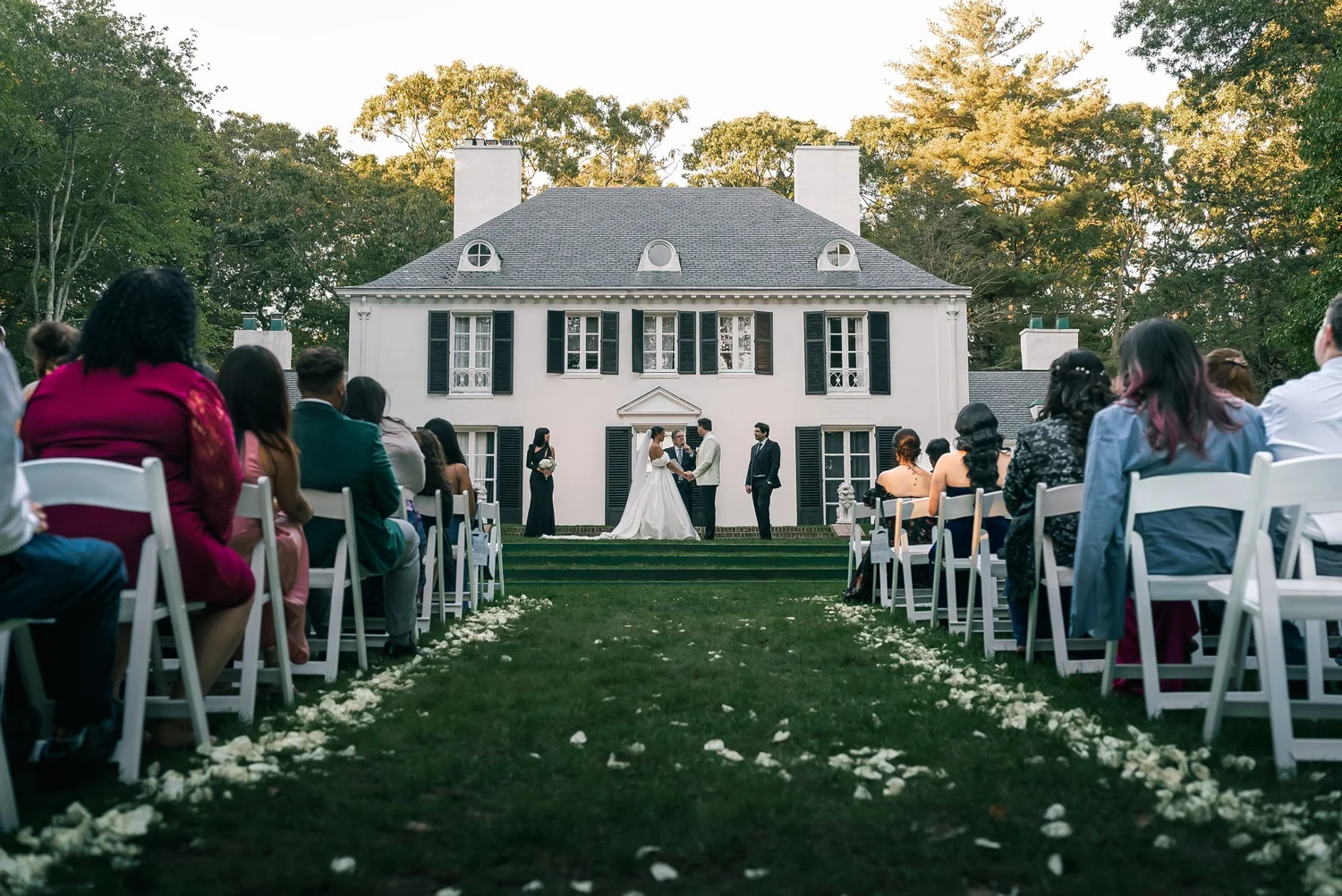 Guests seated on white chairs on either side of a petal-lined aisle watch Patricia and Joshua at the front of a white house with black shutters.