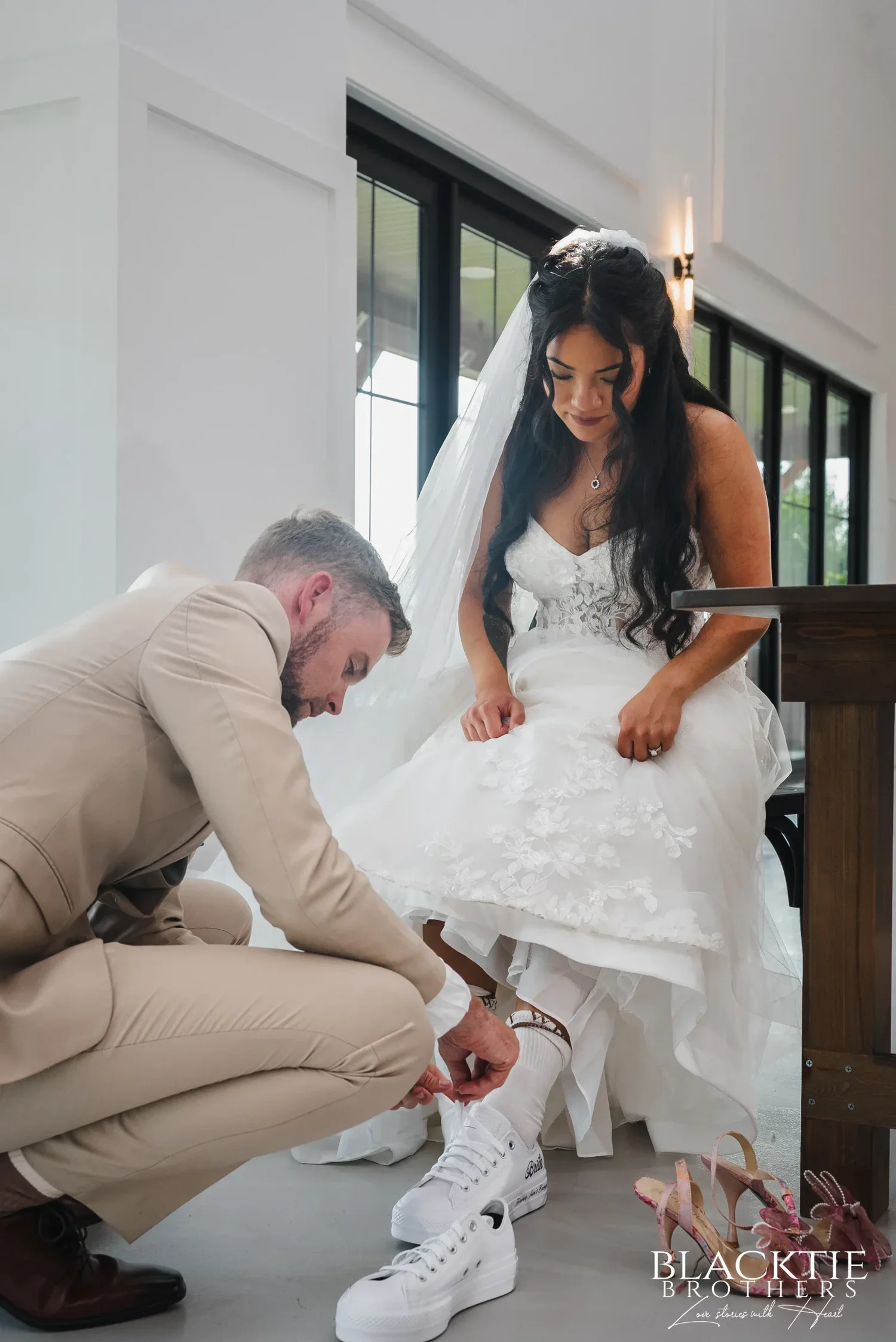 Josh crouching to lace white sneakers onto Carolina's feet while she lifts her dress, her heels on the floor beside them