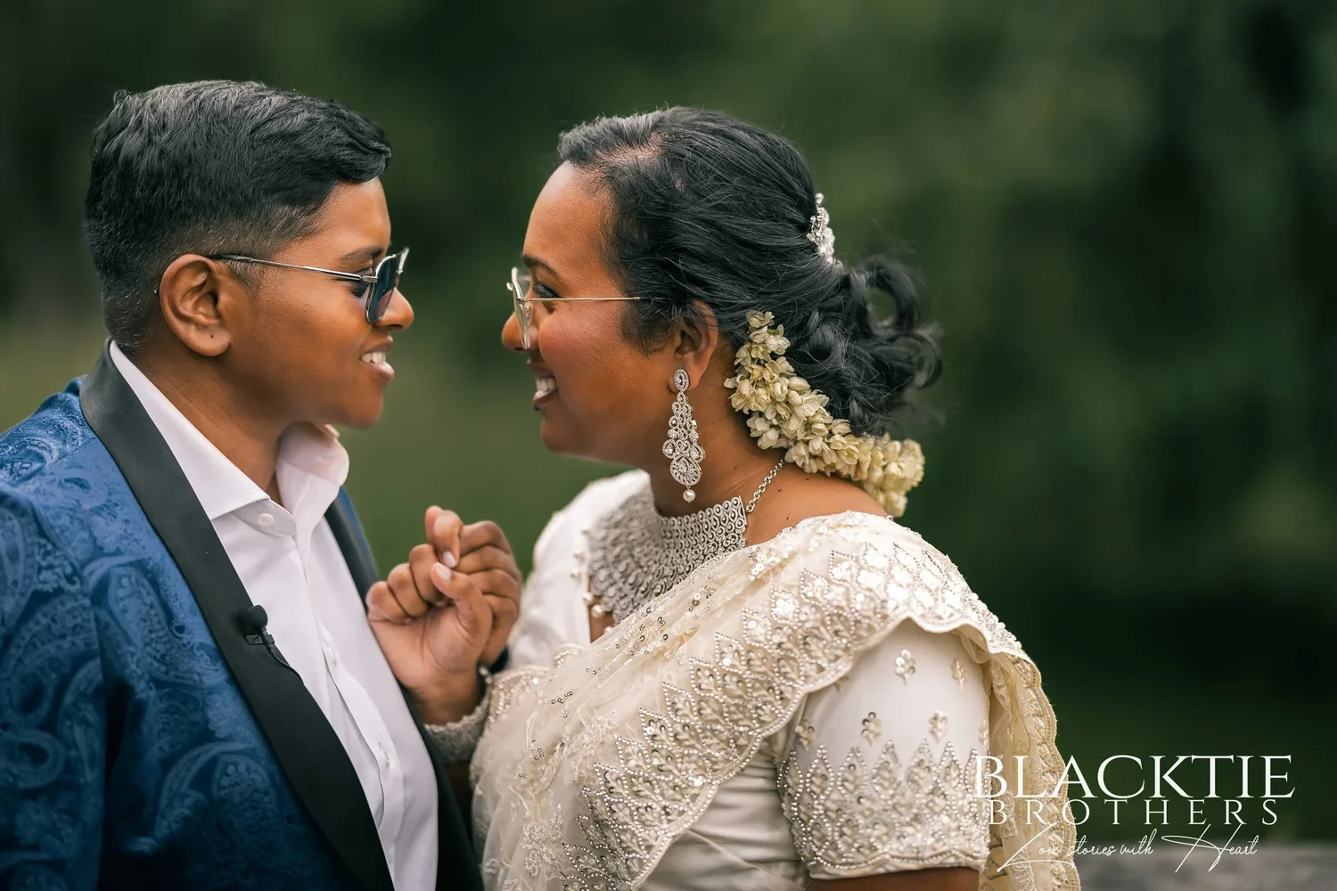 Hima and Alina face to face and laughing in the garden, one in a blue paisley tuxedo jacket and sunglasses, one in an ivory embroidered outfit with white flowers in the hair
