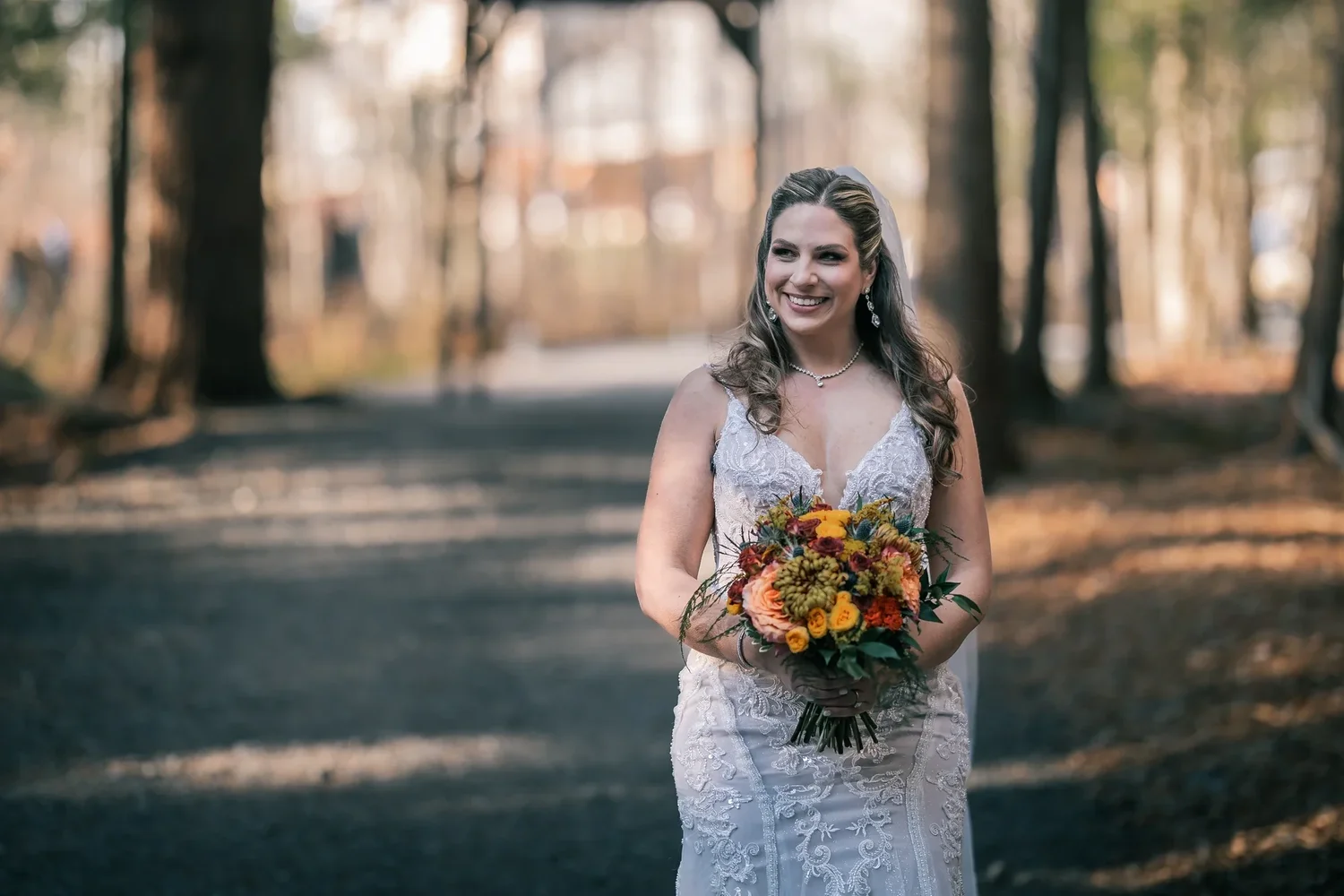 Charissa walking toward the Enchanted Forest ceremony, holding an autumn bouquet of orange and burgundy flowers, the tall pines behind her softly out of focus