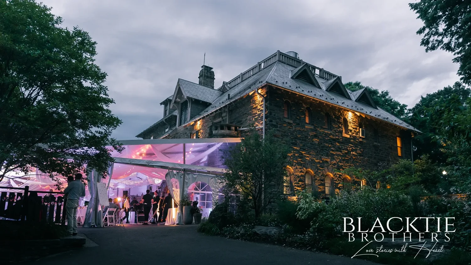 The reception tent lit pink and purple beside the stone carriage house at Morris Arboretum at dusk, guests inside and trees around it