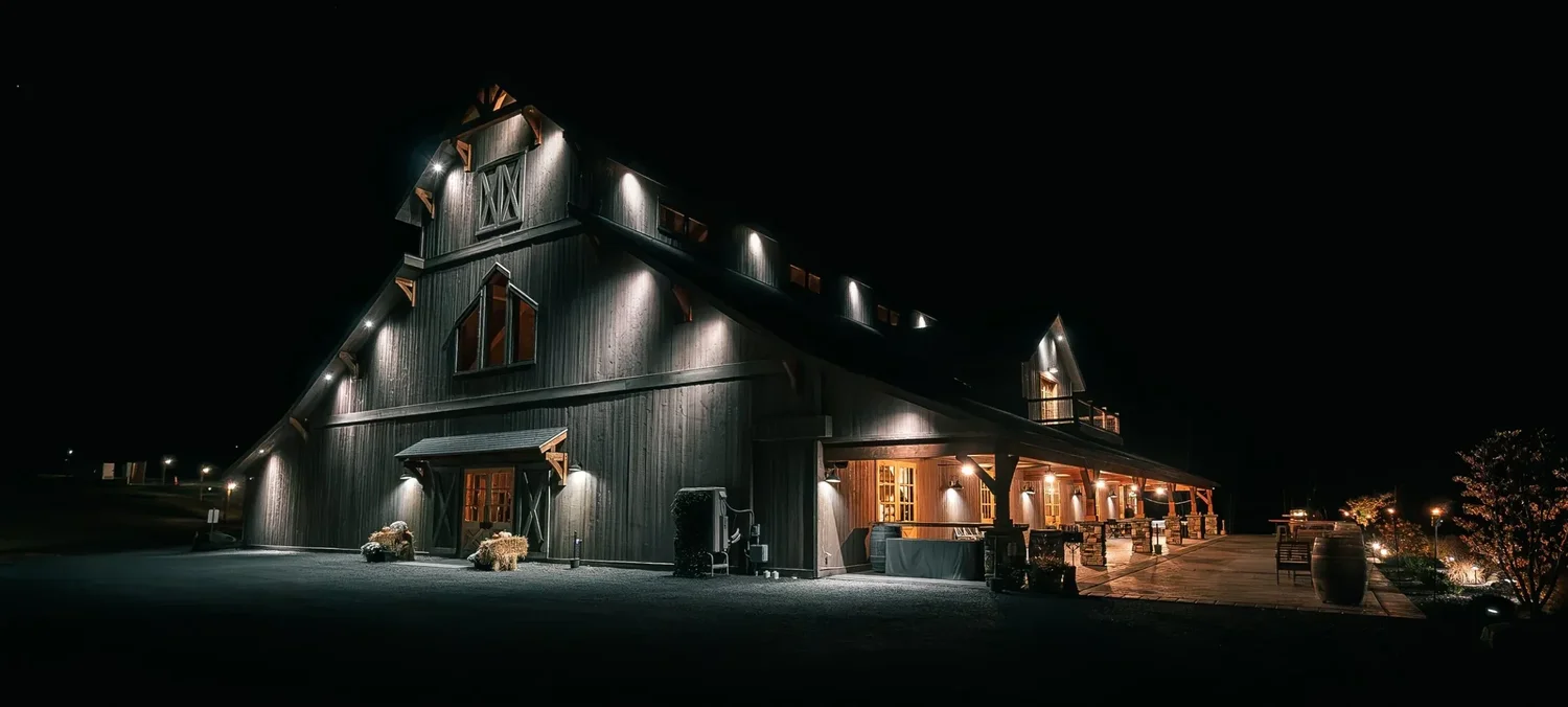 The Barn at Windham Manor at night: board-and-batten walls lit from above, warm light through the doors and windows, and the covered stone patio glowing along one side