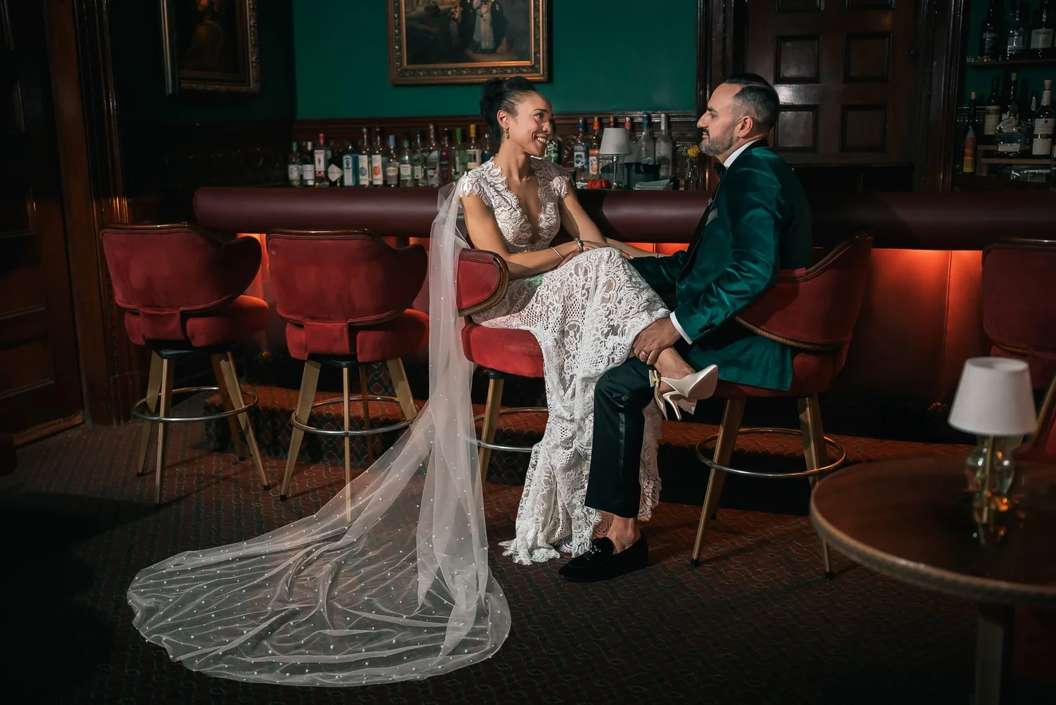 Shannon laughing with Leon on red bar stools at the wood-paneled bar, her long veil spread across the floor