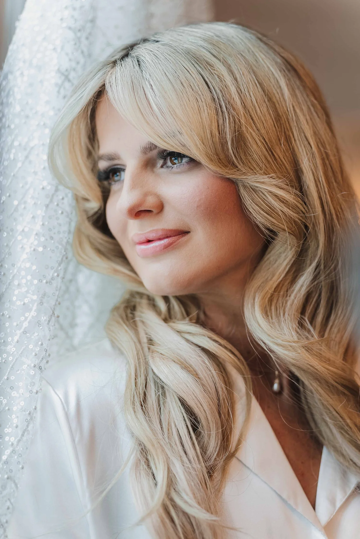 Close portrait of Kari in a white robe by the window, her beaded wedding gown hanging beside her.