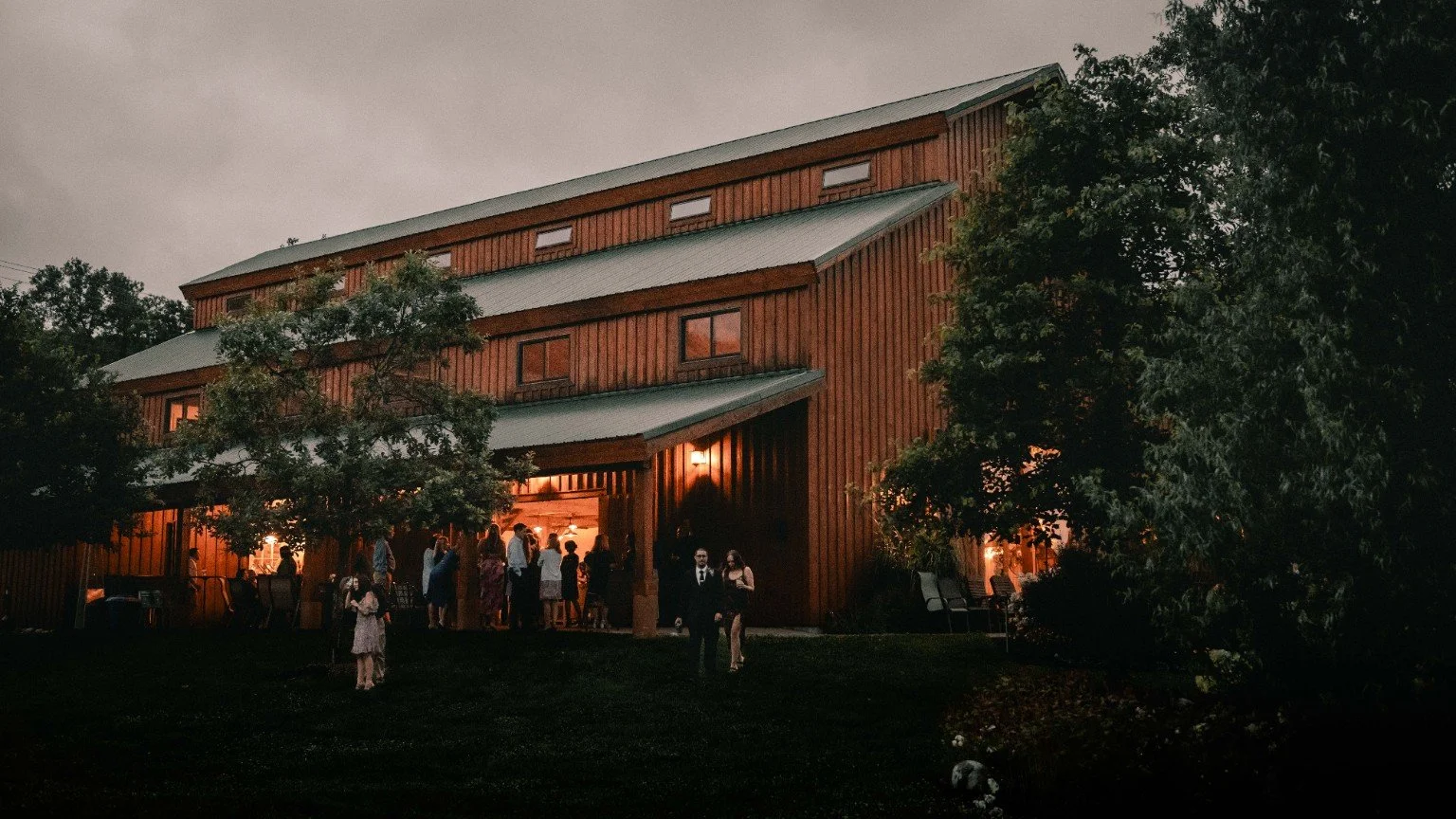 The illuminated timber barn at The Orchards at Rocking Horse Farm, with wedding guests gathered outside at dusk.