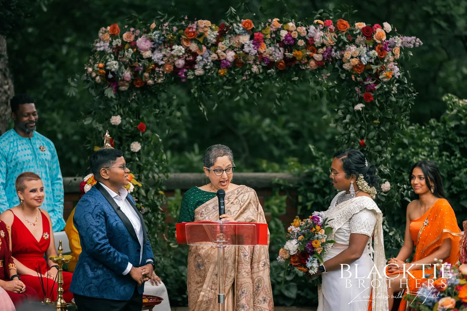 The officiant speaking into a microphone at a red lectern between Hima and Alina under the floral arch on the brick patio, members of the wedding party on either side