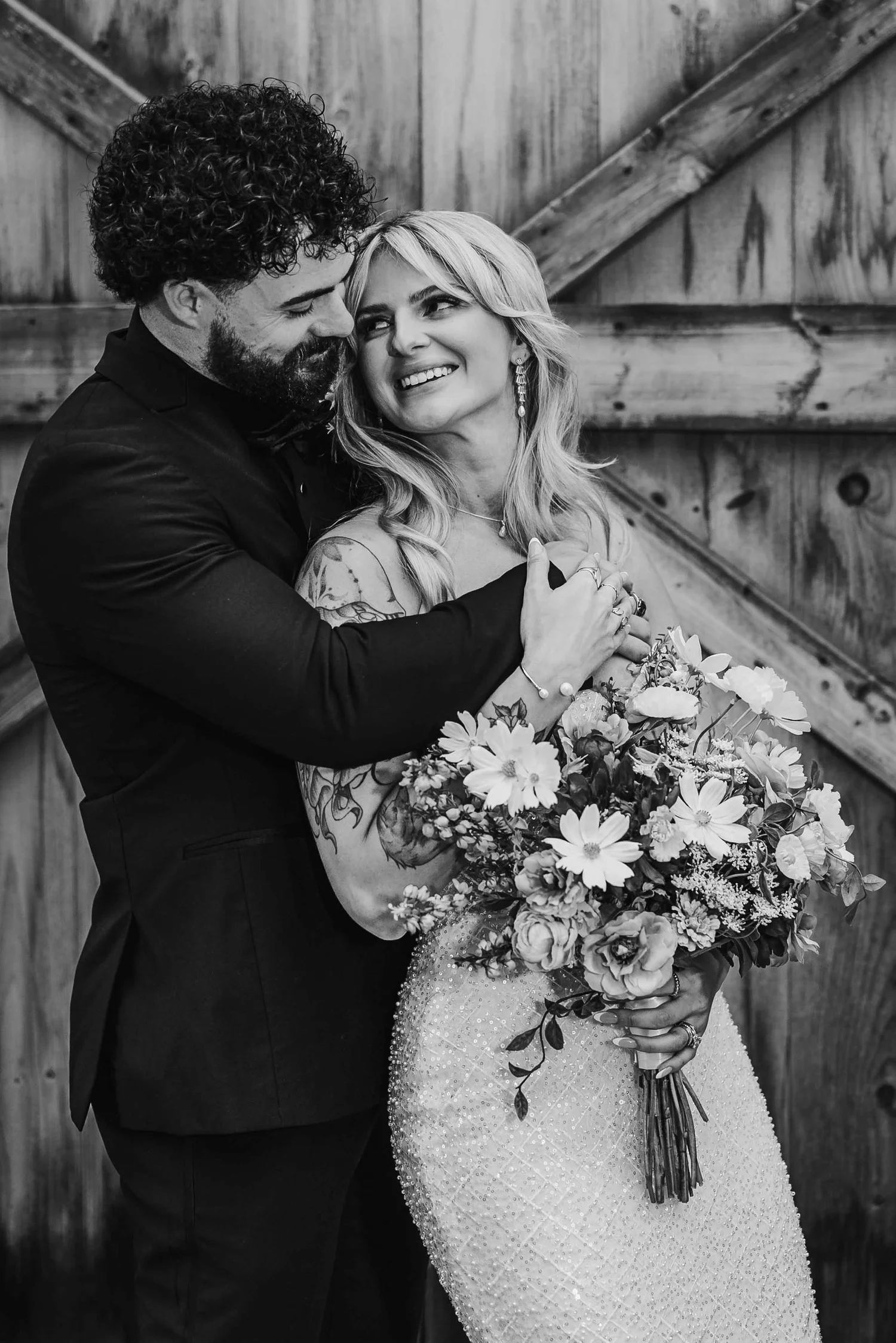 Black and white photograph of Josh embracing Kari from behind against weathered barn doors as she holds her bouquet and smiles.
