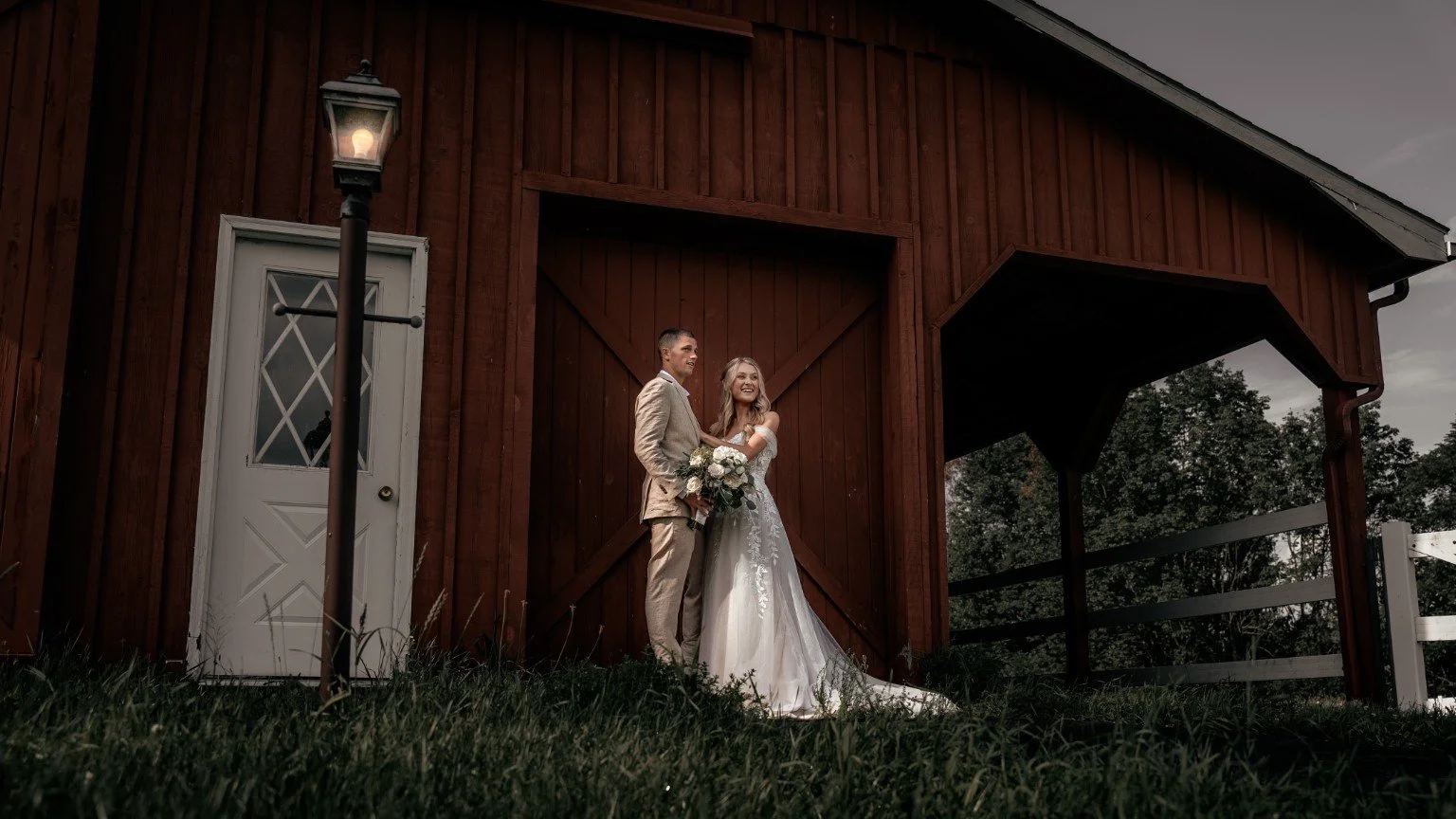 Melissa and Tim smiling together in front of a red barn, with Melissa holding a bouquet.