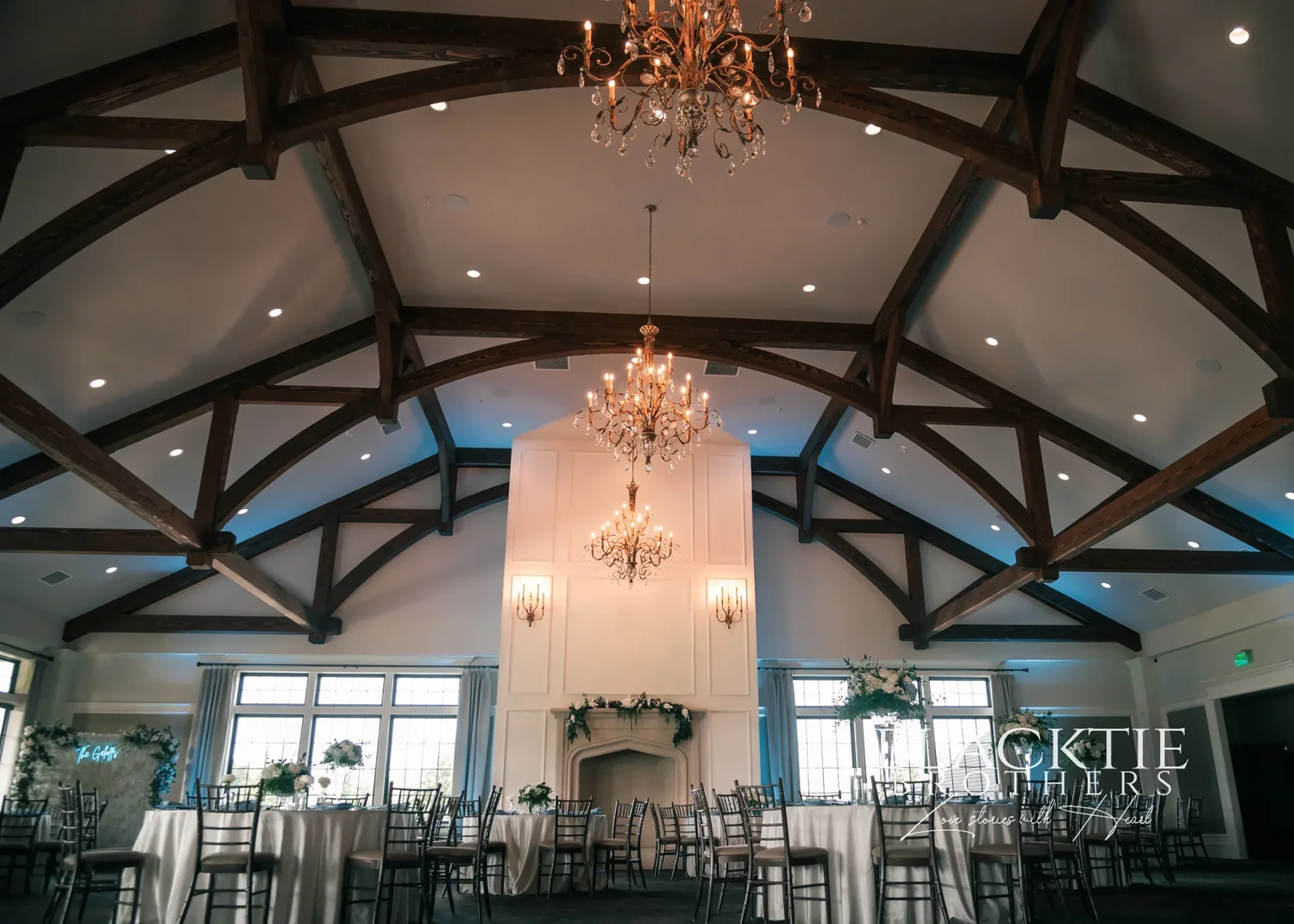 The Grand Ballroom at Timber Banks set for Taylor and Jake's reception: dark timber trusses under a white vaulted ceiling, a crystal chandelier centered above the fireplace, tall windows, round tables with white linens and tall white floral centerpieces