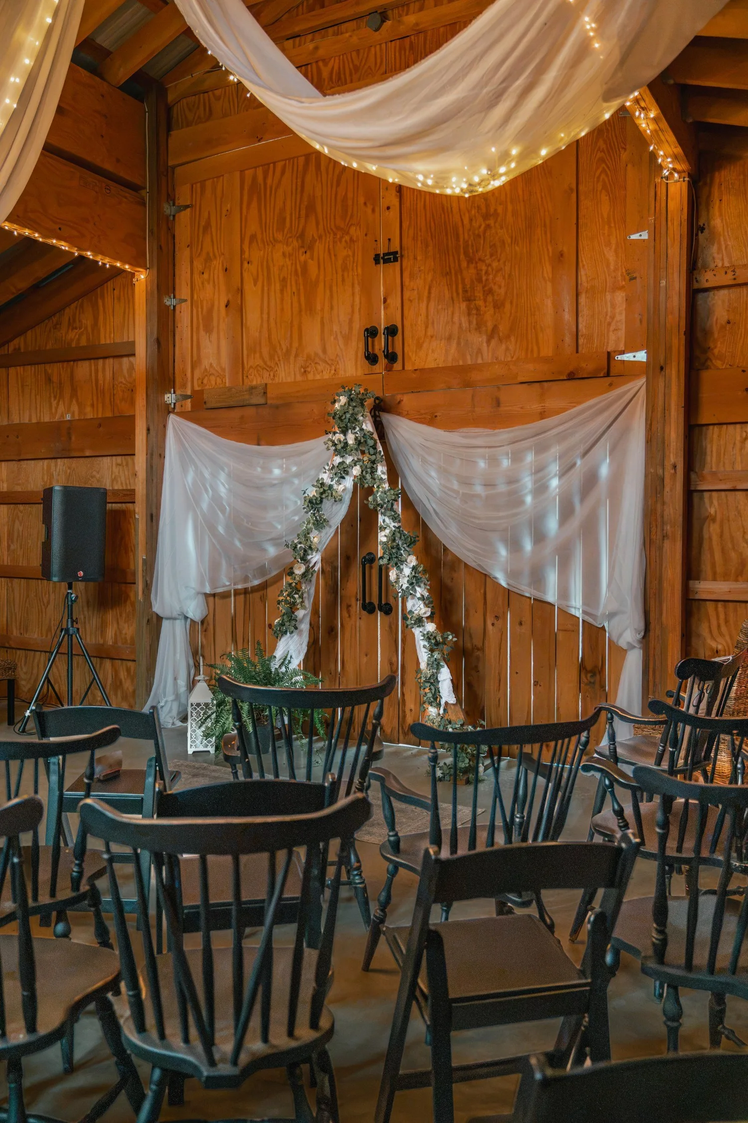 The ceremony setup inside the barn before guests arrive: dark wooden chairs facing a white draped backdrop and a greenery arch, with string lights along the beams