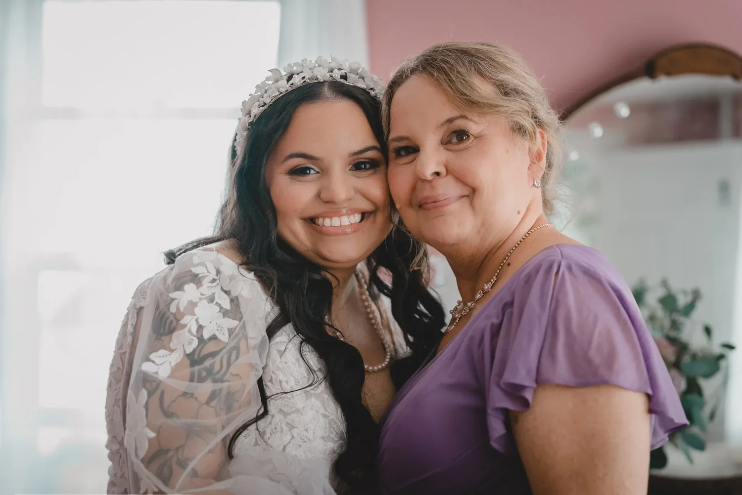 Jessica in a floral tiara, cheek to cheek with a woman in a lilac dress, both smiling while getting ready