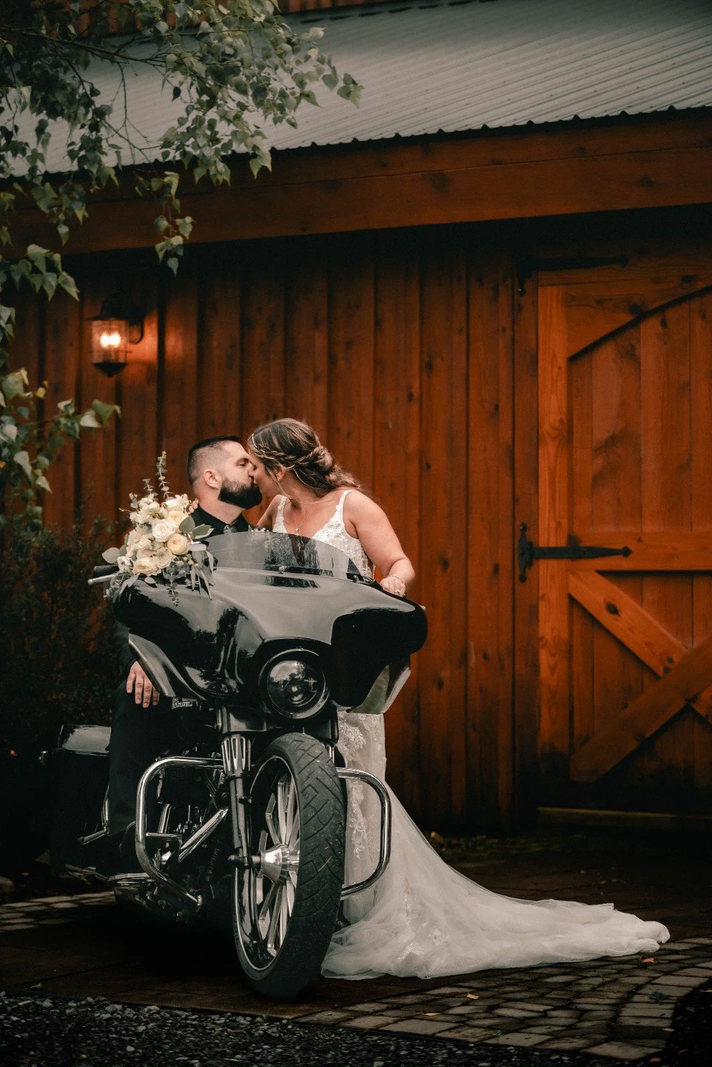Joelle and Louie kiss beside a black motorcycle in front of the wooden barn doors.