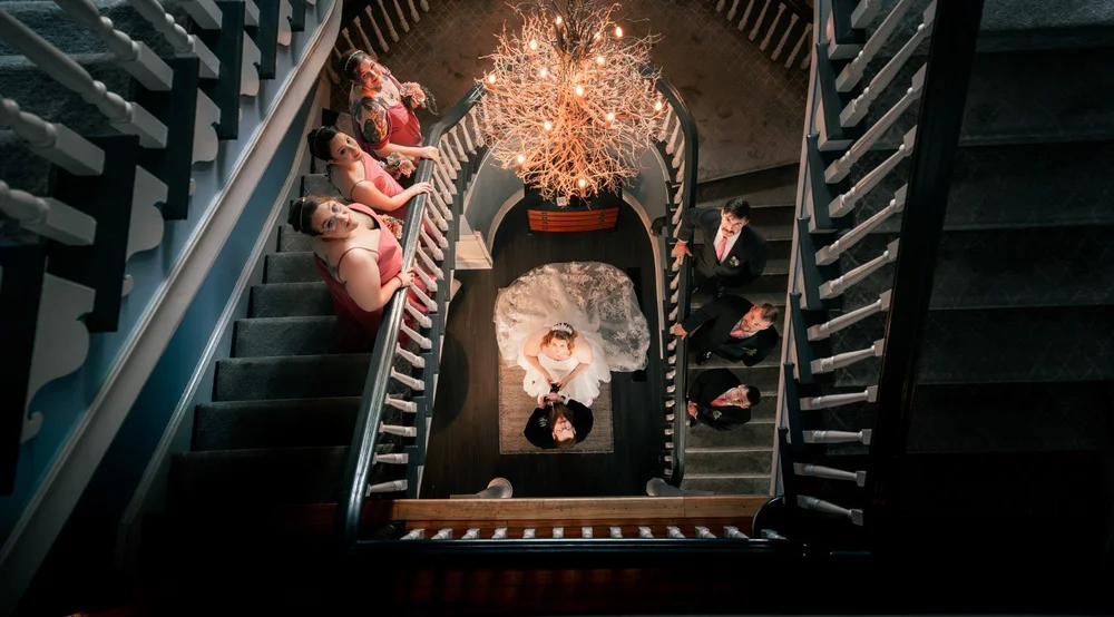 Overhead view of a wedding couple and attendants arranged around a staircase beneath a chandelier.