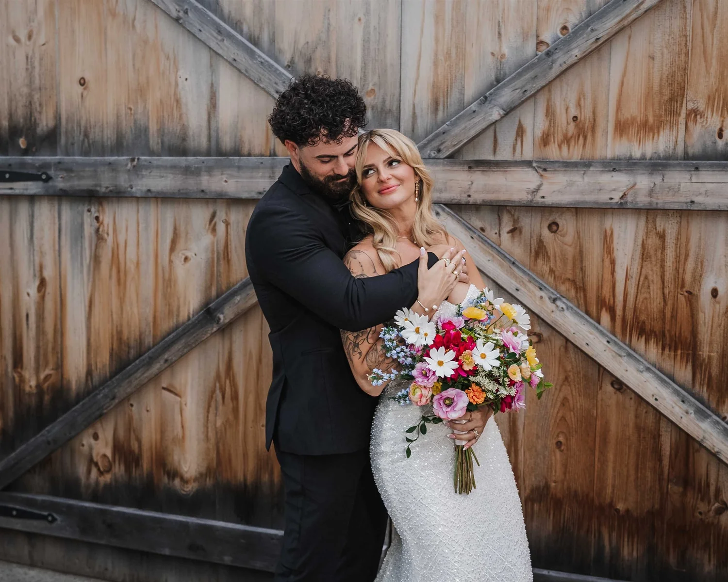 Josh embraces Kari from behind as she holds her colorful bouquet in front of wooden barn doors.