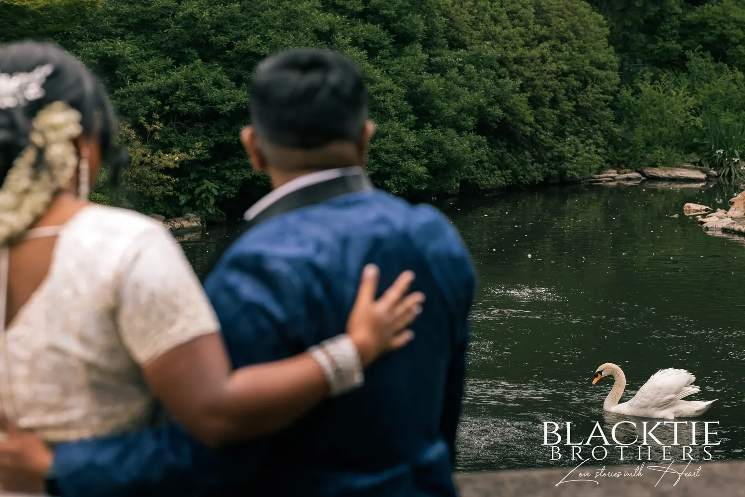 Hima and Alina seen from behind with their arms around each other at the edge of a pond, a white swan gliding past on the water