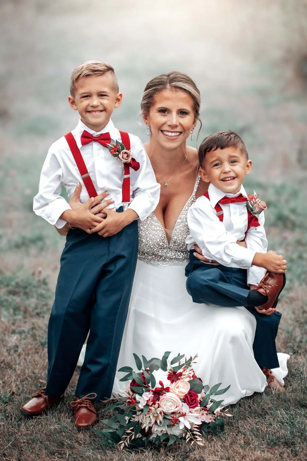 A bride sits outdoors with two smiling boys in red bow ties and suspenders, with a bouquet at their feet.