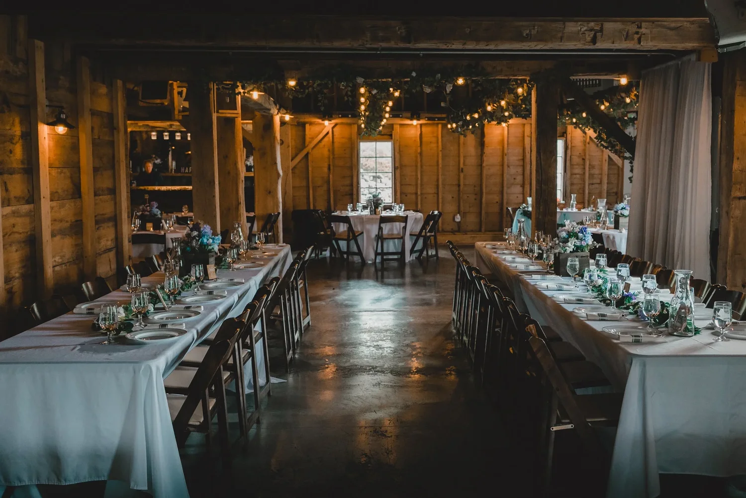 Long dinner tables set with white linens, greenery and glassware in the Grand Barn Hall at Knotting Hill Farm