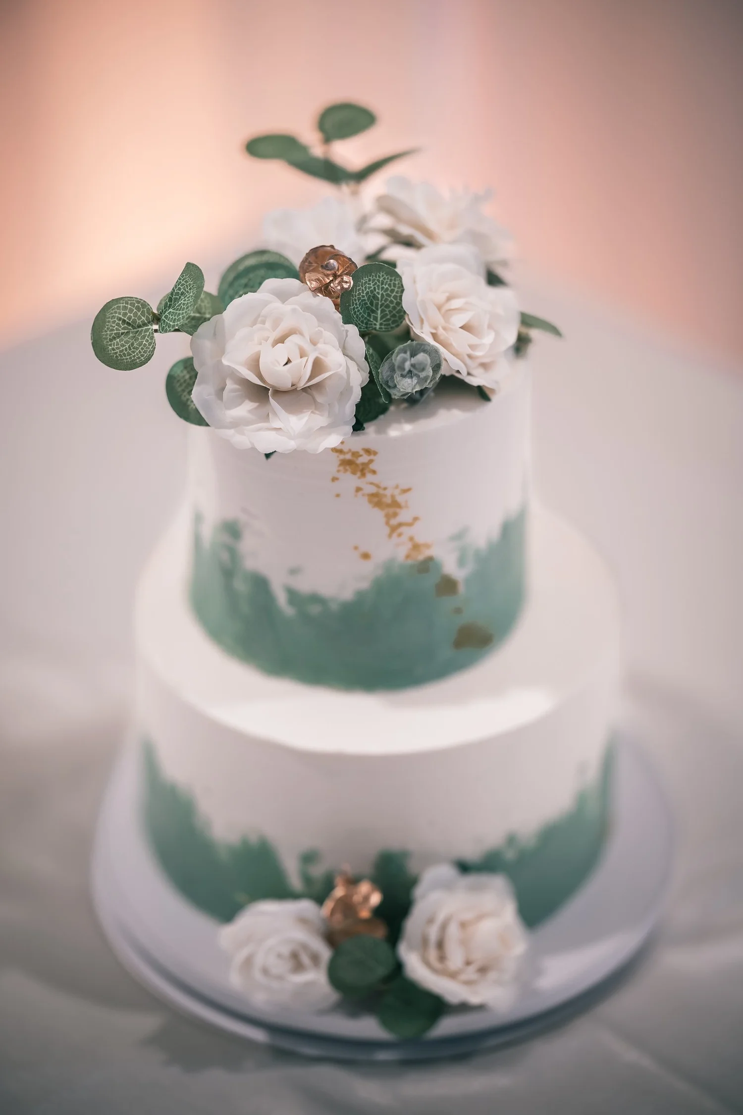 A two-tier white wedding cake brushed with green and gold leaf, topped with white roses and eucalyptus.