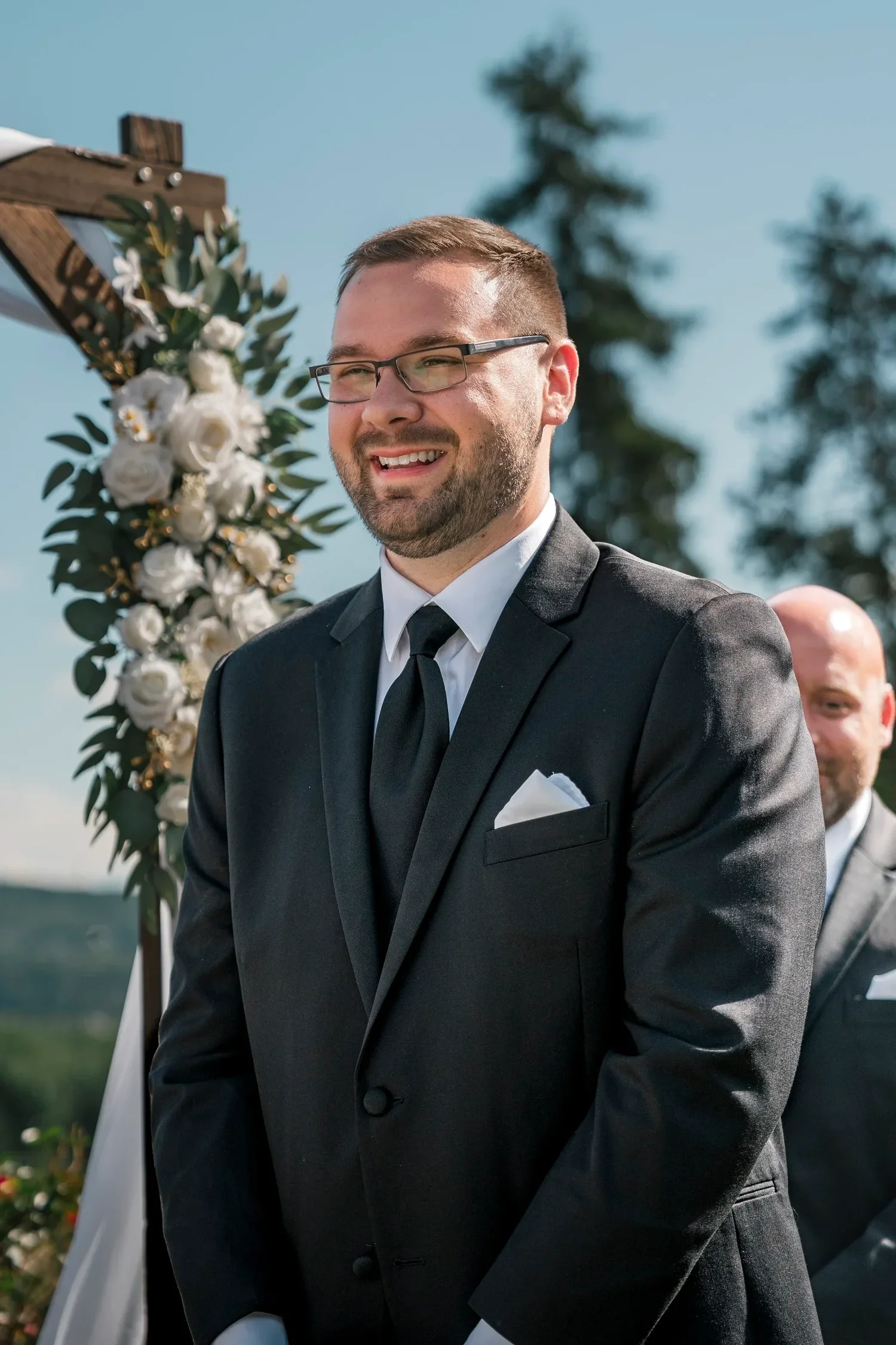 Zach smiling beside the floral arbor during the ceremony at Traditions at the Glen