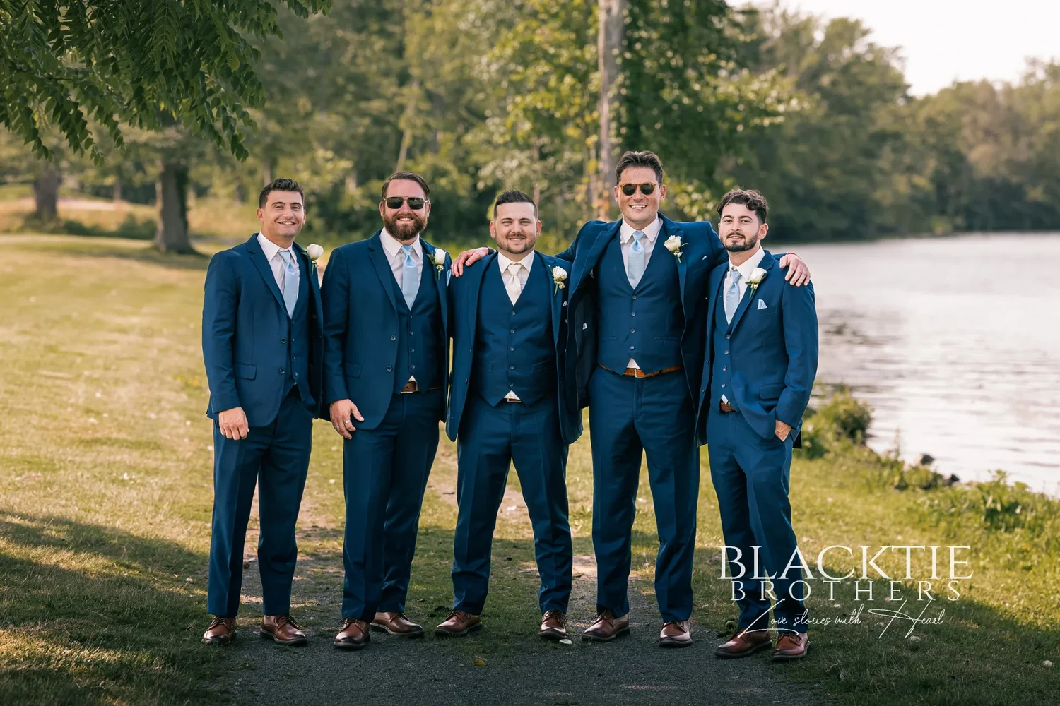 Jake and his four groomsmen in navy three-piece suits standing arm in arm on the grass beside the Seneca River at Timber Banks, trees and the water behind them