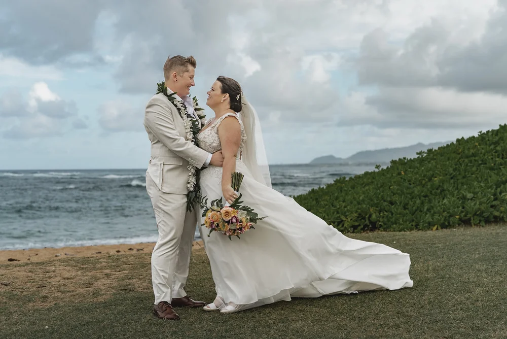 A couple in wedding attire holding a bouquet together beside the ocean in Hawaii as the dress moves in the breeze