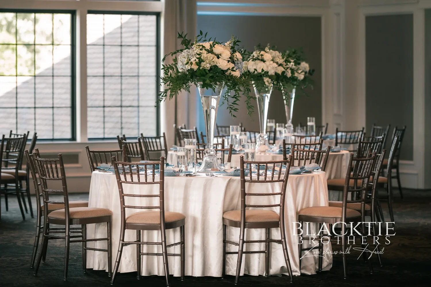 A round reception table at Timber Banks set with white linens, gold chairs and a tall white and green floral centerpiece, tall windows and the ballroom beyond