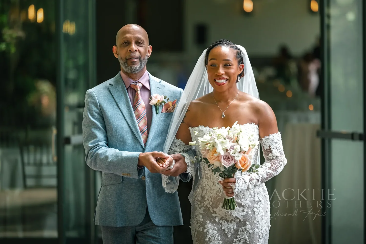 The bride in her lace gown and veil, holding her bouquet, walking arm in arm with a man in a blue suit through glass doors