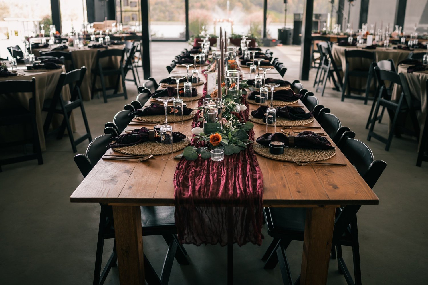 A long wooden farm table set with dark plates, a burgundy runner and greenery inside the pavilion
