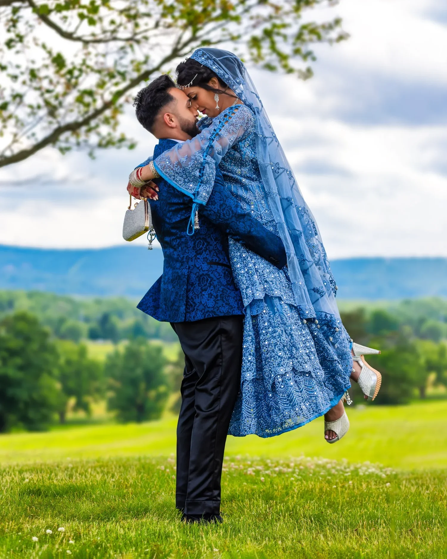 A groom in a blue brocade jacket lifting his bride, in a pale blue embroidered gown and veil, beneath a tree on the hill at Traditions at the Glen with the ridge behind them
