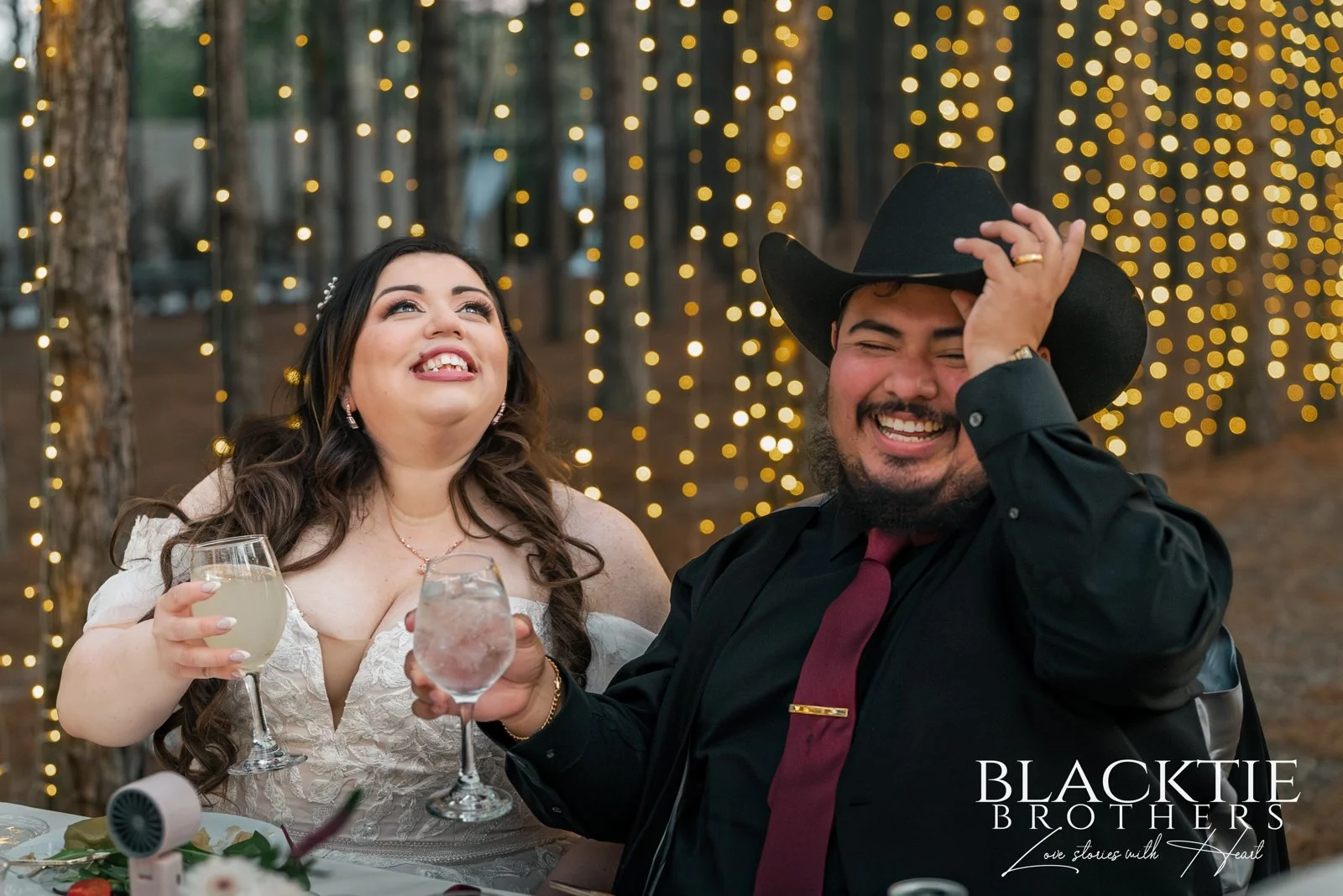 Alyssa and Alberto laugh together with glasses in hand beneath warm string lights at their Pinery wedding.