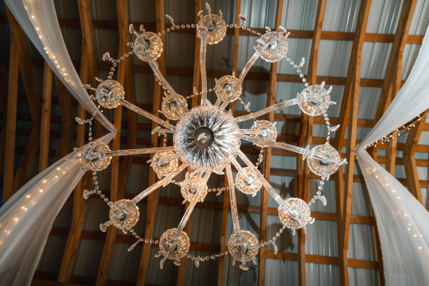 Looking straight up at the crystal chandelier in the Black Barn, white fabric and string lights draped across the wooden rafters