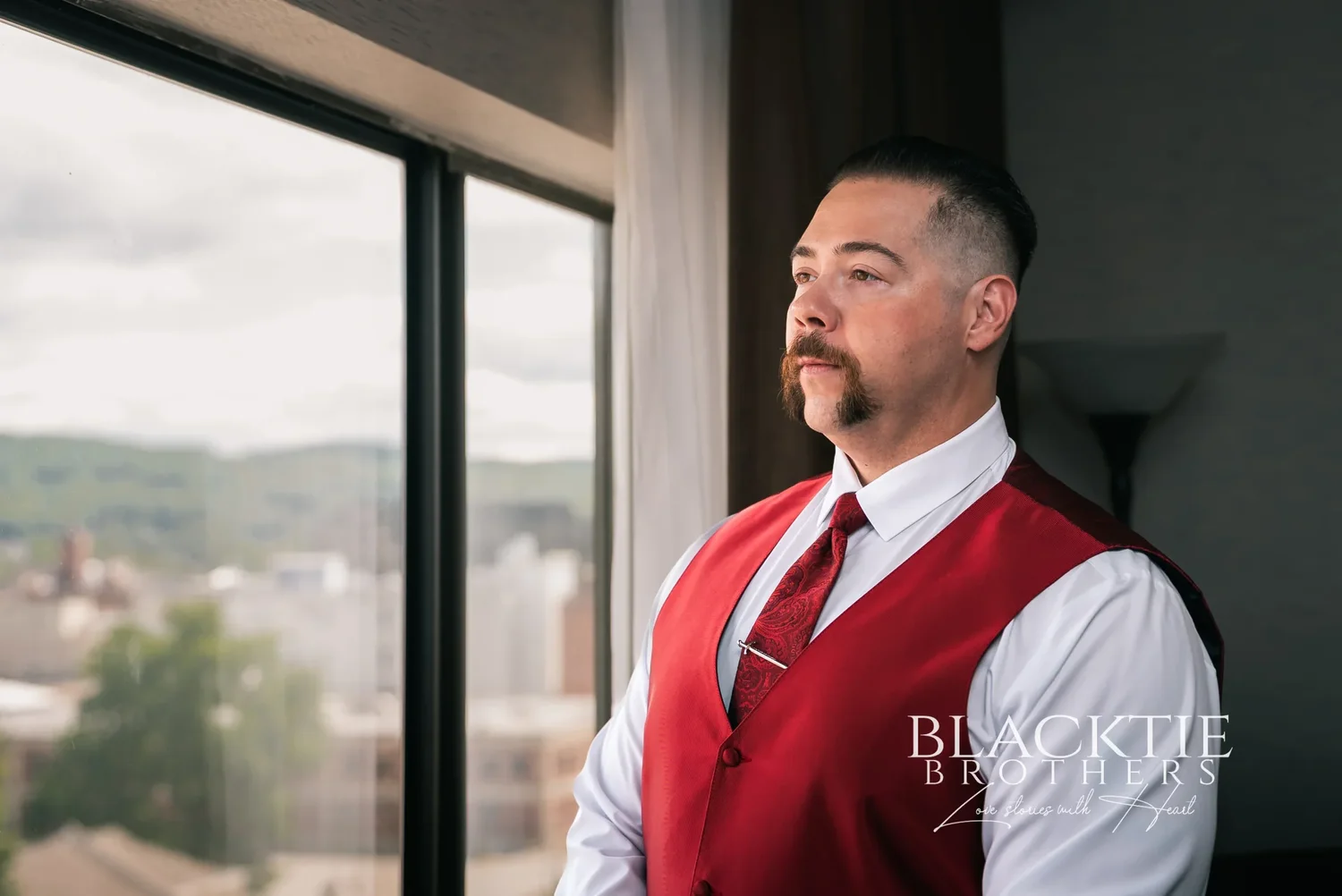 Dante in a red vest and tie standing beside a hotel room window at the DoubleTree by Hilton Binghamton, looking out over downtown and the hills