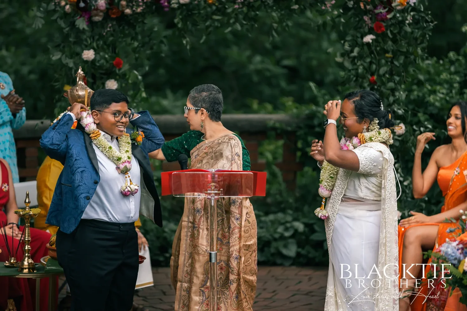 Hima and Alina wearing thick flower garlands during the ceremony, the officiant between them at the red lectern