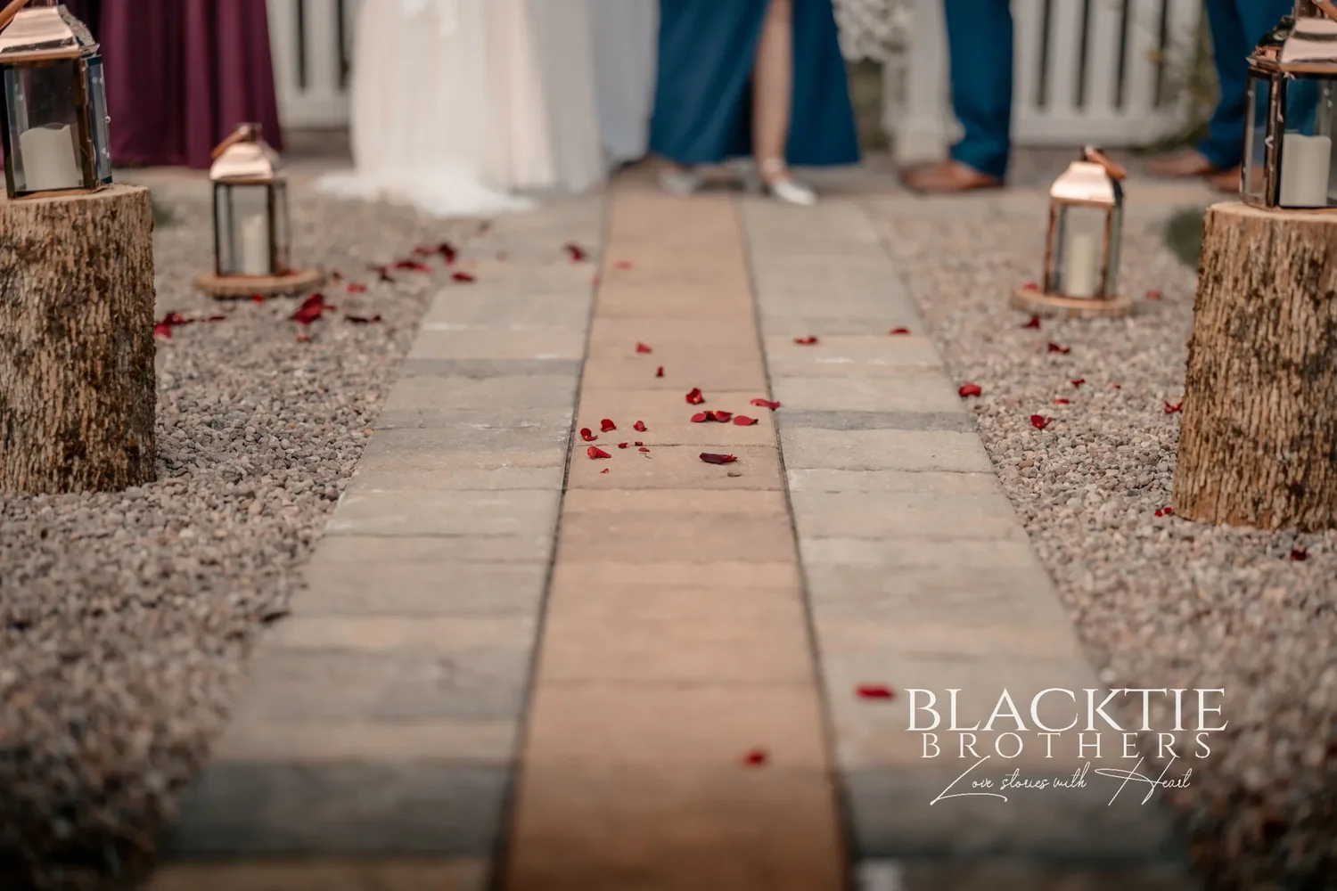 The empty ceremony aisle at The Mason Jar before guests arrive: a paver path edged with gravel, copper lanterns on log rounds and red rose petals