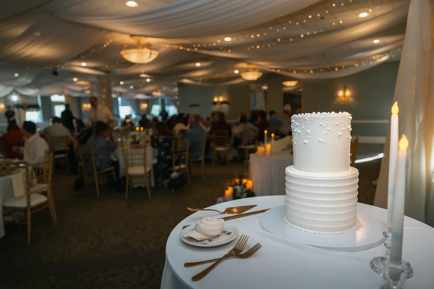 The reception room at Traditions at the Glen with a draped ceiling and string lights, a white wedding cake in the foreground and guests at their tables