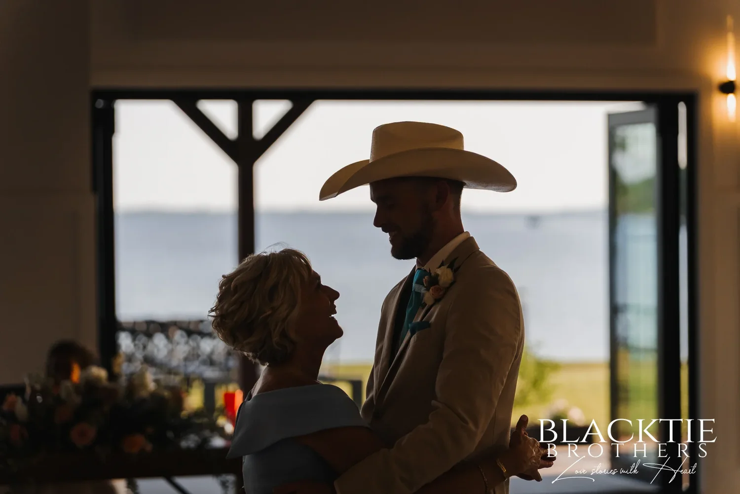 A dance in silhouette in front of the open glass doors, Josh in a cowboy hat, the lake bright behind them