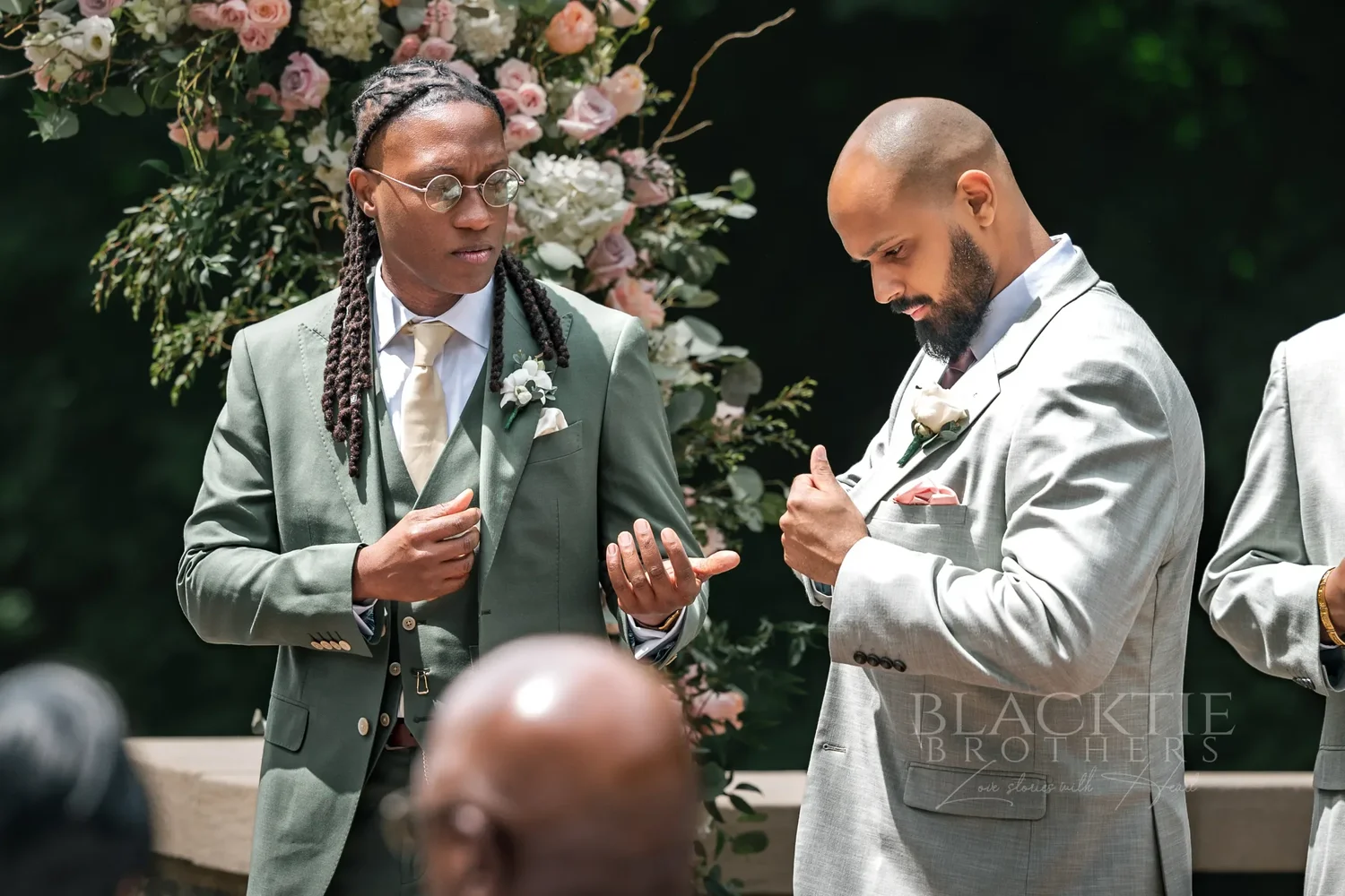 The groom in a sage-green suit and a groomsman in a light gray suit adjusting their jackets and cuffs beside a floral arch