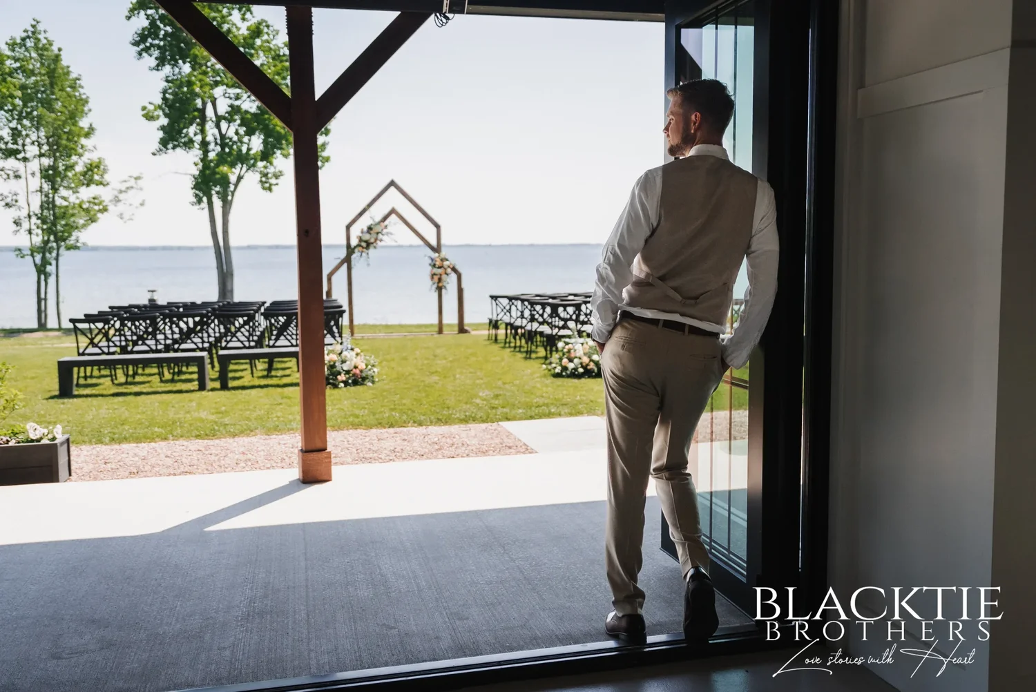 Josh standing in the open glass doors of the event center, looking out at the empty ceremony chairs and the wooden arch on the lawn by the water