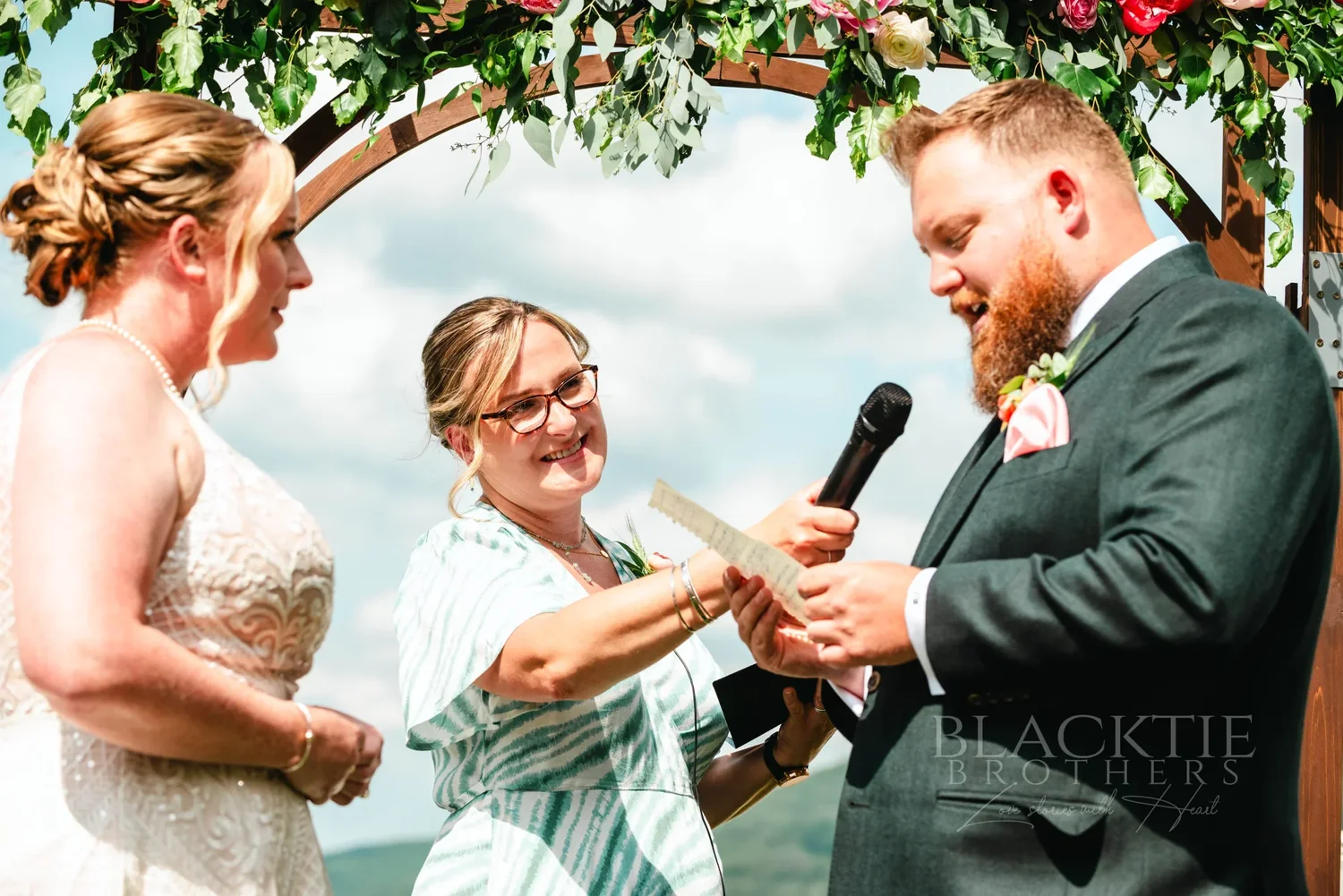 Amanda and Chris laughing with their officiant under the floral arbor at The Otesaga