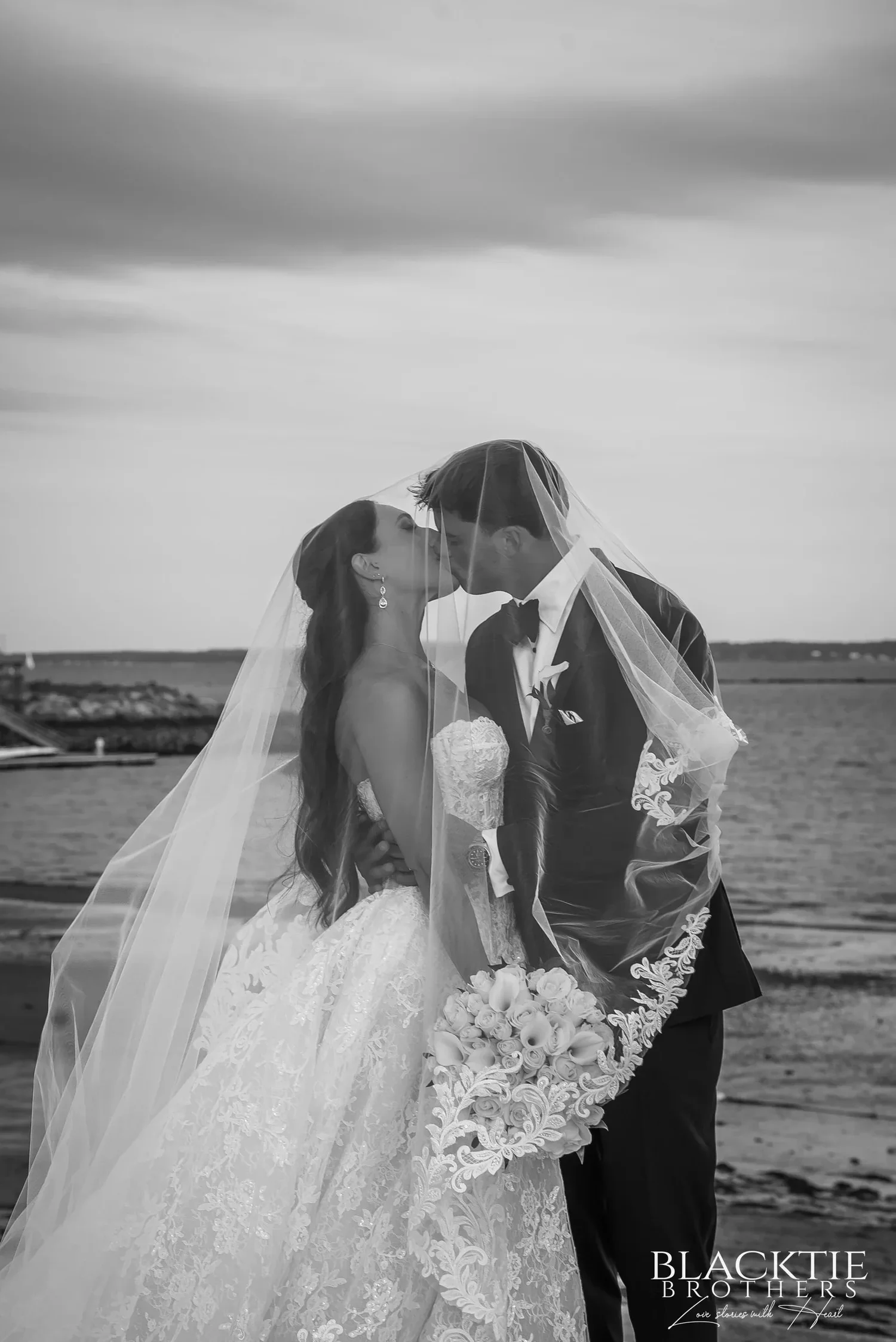 Black and white photograph of Leah and Nicholas kissing under her veil at the water's edge, her bouquet of white roses between them