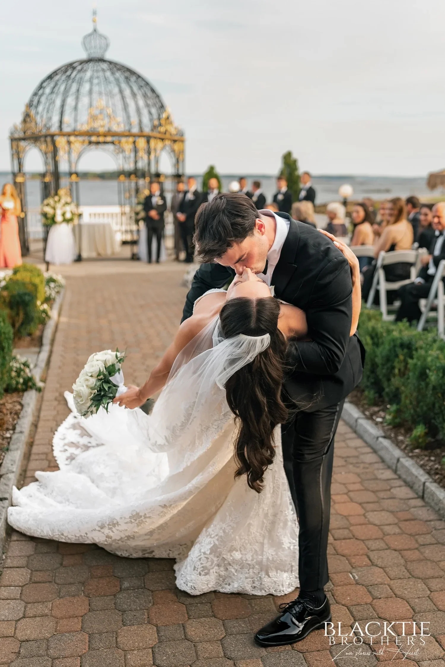 Nicholas dipping Leah into a kiss on the brick aisle after the ceremony, her veil and train flowing, guests and the domed gazebo behind them by the water