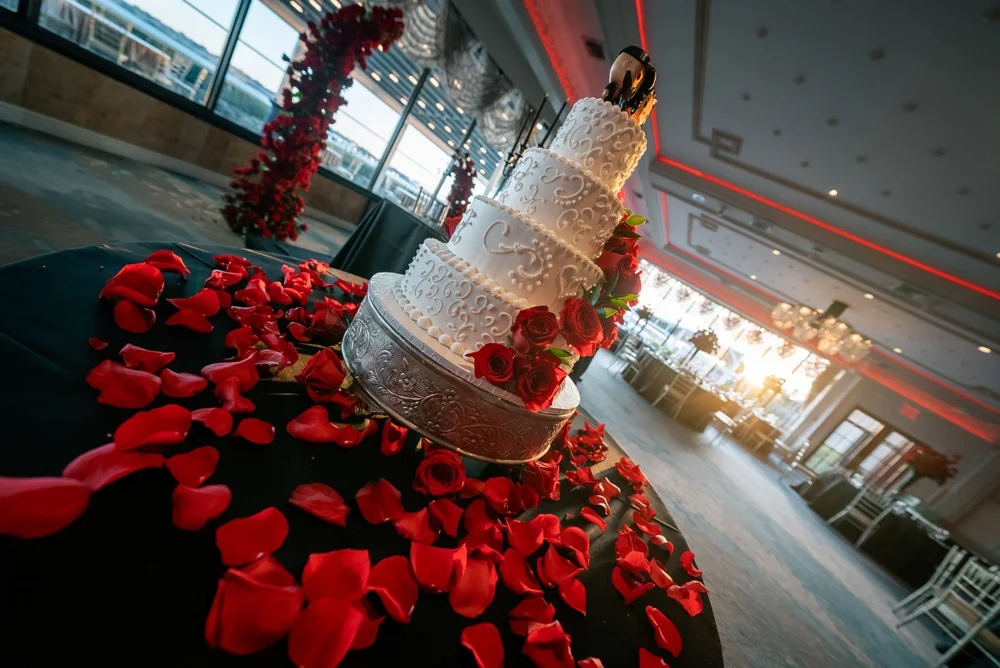 White tiered wedding cake decorated with red roses on a table scattered with red petals.