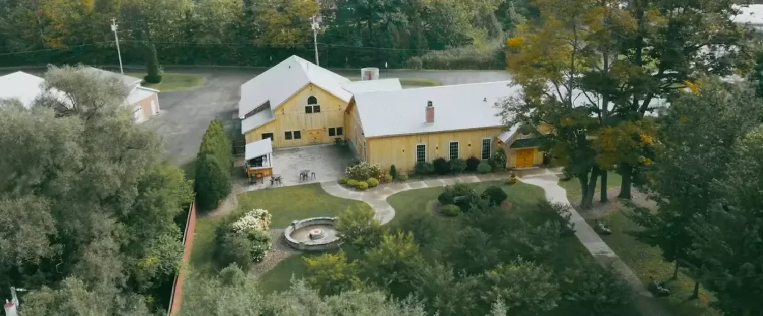 Aerial view of The Cannery: a long yellow board-and-batten building with a metal roof and a round gable window, curved paths and a stone fountain in the garden