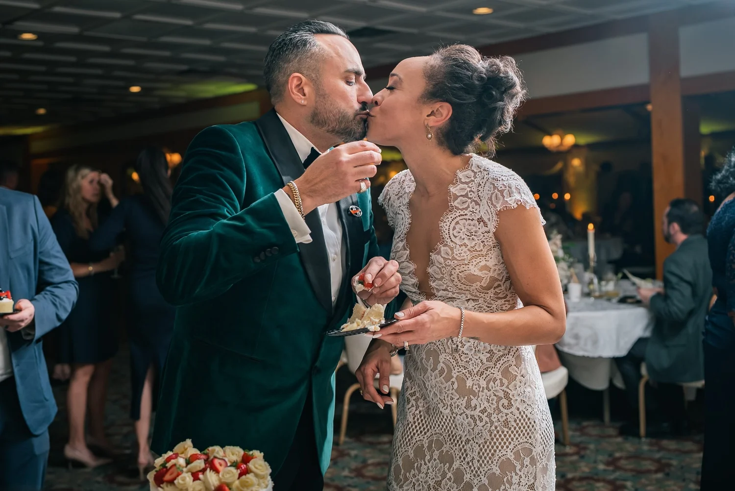 Leon and Shannon sharing a kiss mid-bite at the cake, with guests at their tables behind them