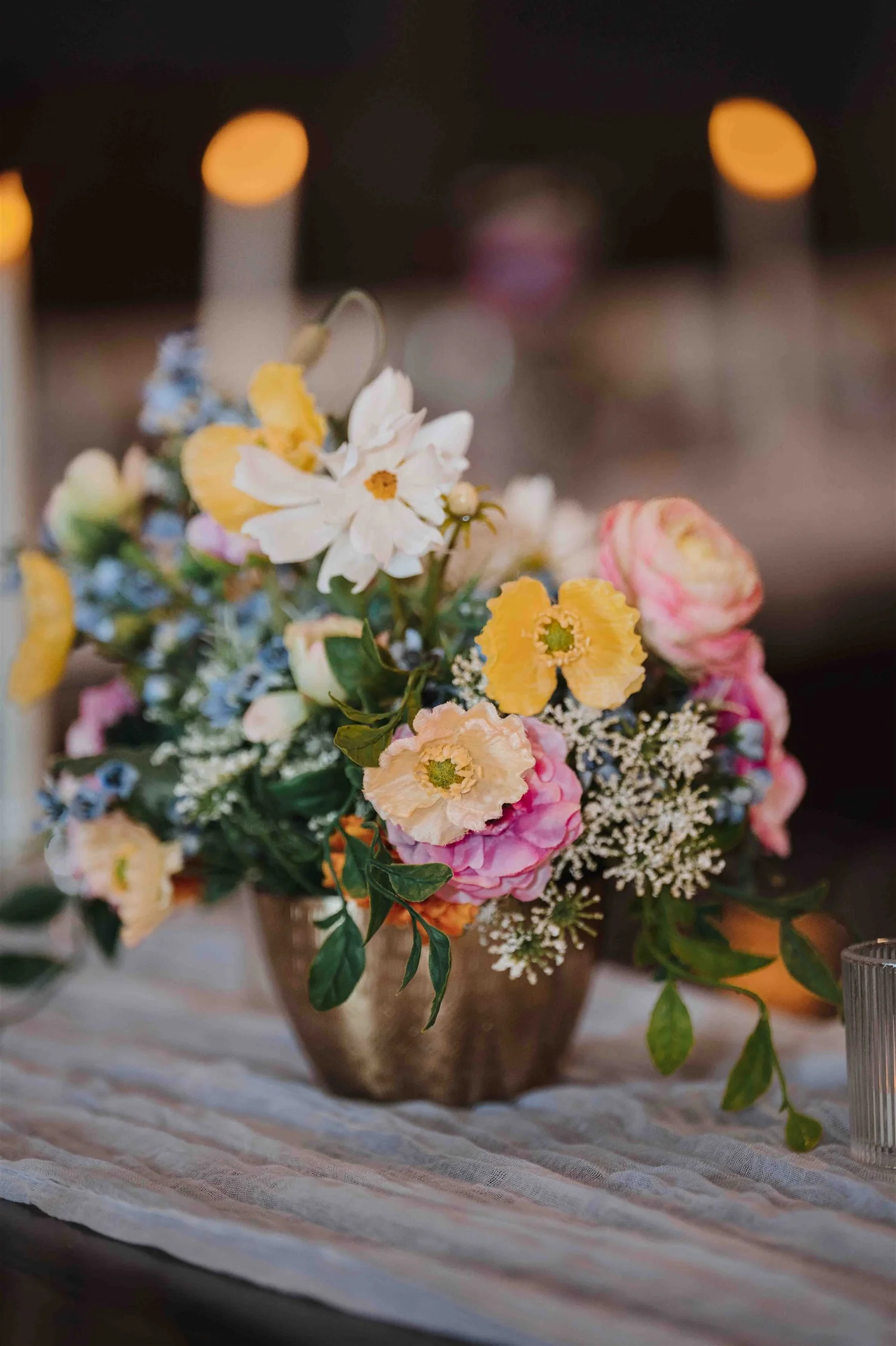 Pink, yellow, white and blue flowers in a brass-colored vase on a softly draped reception table.