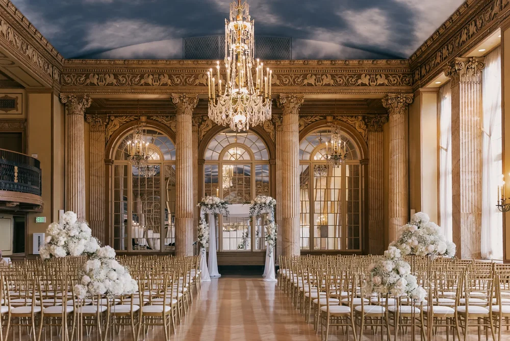 Wedding ceremony set with white flowers and gold chairs beneath a crystal chandelier and painted sky ceiling at Marriott Syracuse Downtown