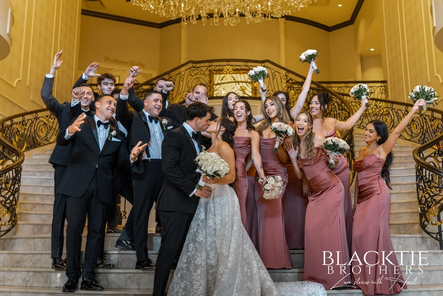The wedding party cheering on the marble staircase, bridesmaids holding white bouquets in the air and groomsmen raising their arms, as Leah and Nicholas kiss in the middle